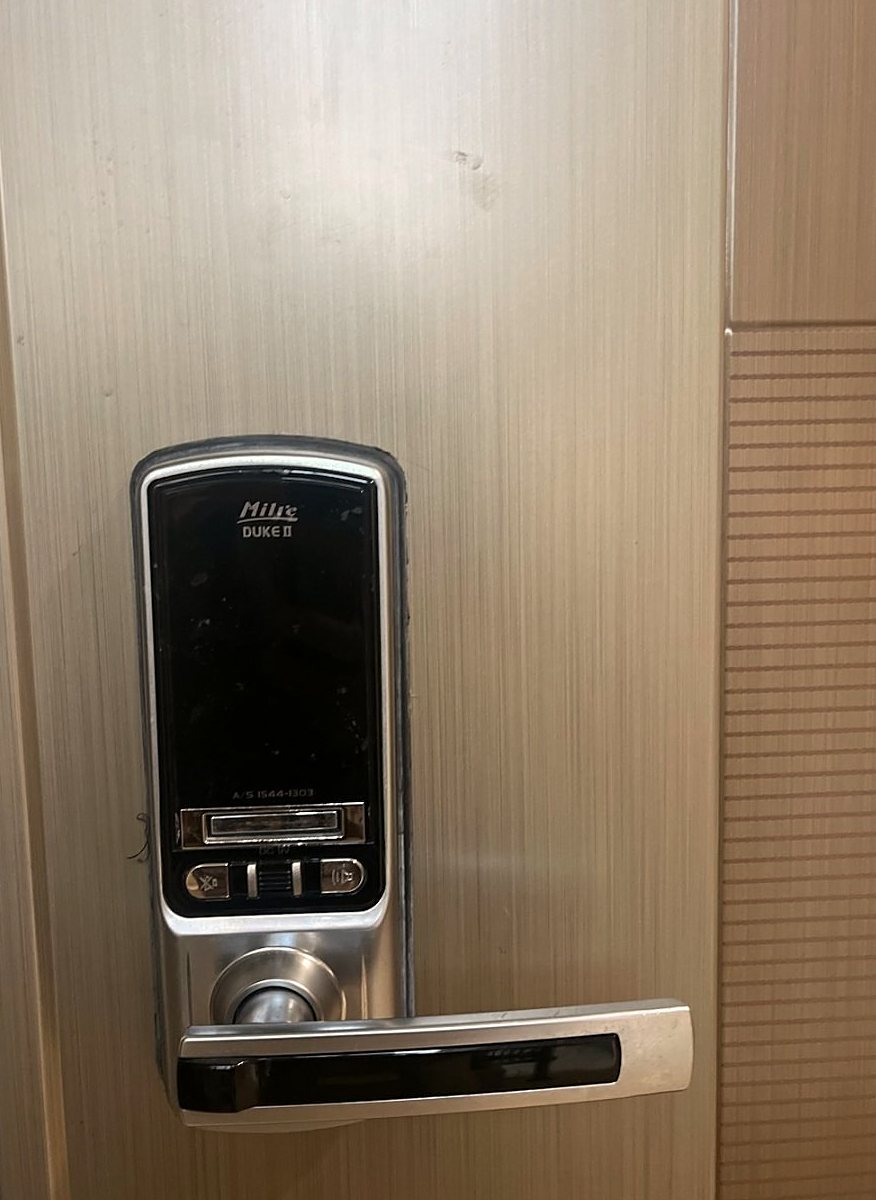 안산시 부곡동 출장 — 결무늬 금속빛 문에 달린 검은 화면의 도어락과 아래쪽 덮개, 버튼 세 개, 은색 손잡이